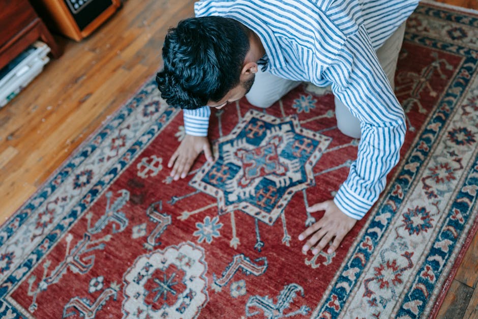 A person dressed in a blue and white striped shirt is kneeling on a patterned red and blue Persian-style rug inside a room with wooden flooring. They are inspecting or cleaning the detailed design, wh