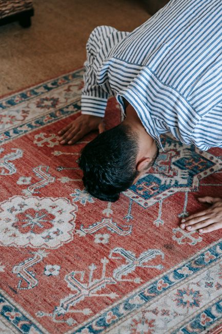 A person dressed in a blue and white striped shirt is kneeling on a patterned red and blue Persian-style rug inside a room with wooden flooring. They are inspecting or cleaning the detailed design, wh