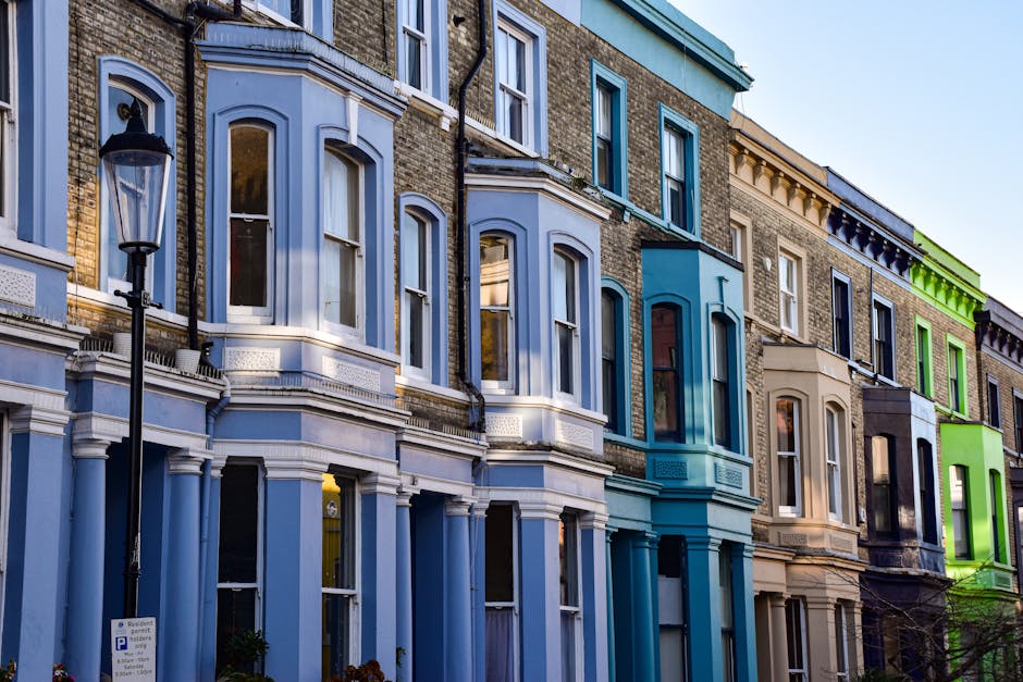 A row of multi-story residential buildings with Victorian-style architecture, featuring bay windows and decorative moldings. The buildings are painted in various pastel shades, including light blue, b