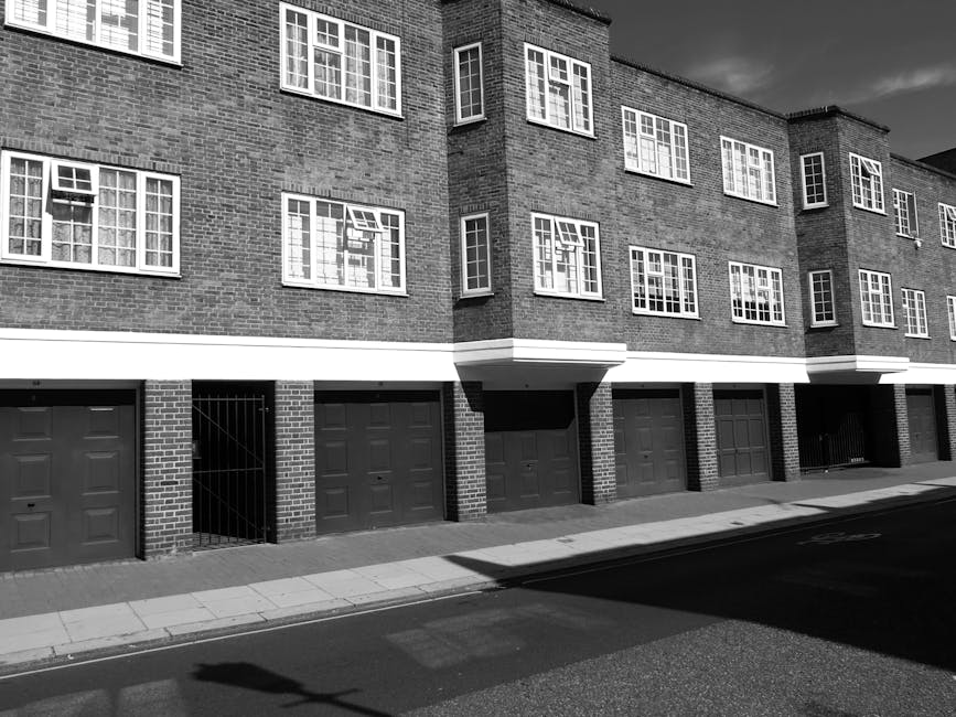 Image of a multi-story residential or commercial building with a brick facade, featuring large arched windows on each floor, set against a pale sky. The building has a prominent sign that reads 'DEVON